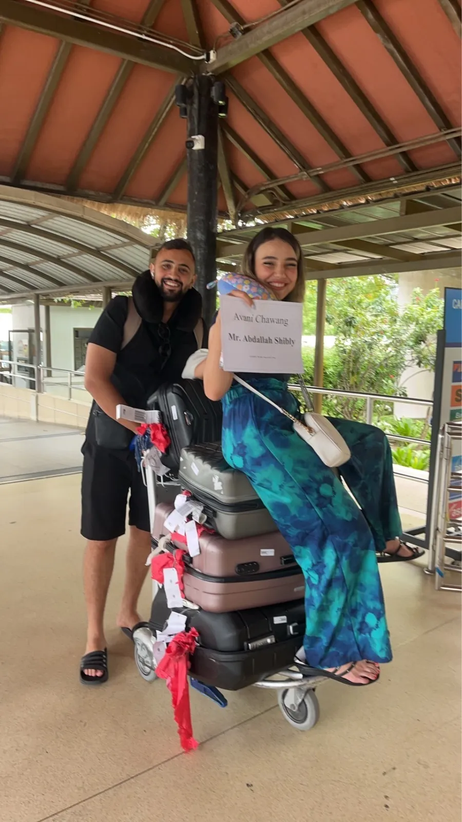 ลูกค้าคู่หนึ่งถือป้ายต้อนรับ ยืนกับรถเข็นกระเป๋าที่สนามบินสมุย